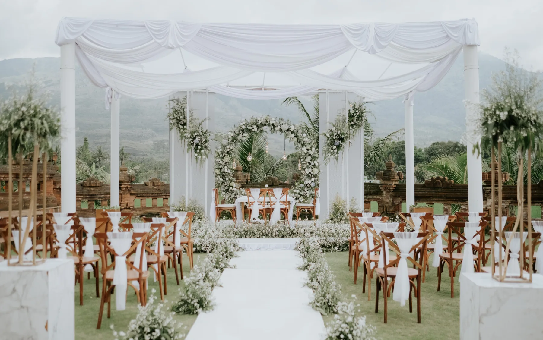 Dekorasi pernikahan Rancabango: floral arch, kain putih, dan kursi kayu dengan latar gunung